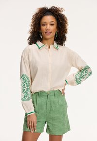 Camicia button-up color crema con accenti ricamati verdi su colletto e maniche, abbinata a pantaloni corti verdi a trama.