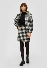 Zwart-wit houndstooth jasje gecombineerd met een bijpassende rok. Het jasje heeft een cropped ontwerp en een rits aan de voorkant. Gladde textuur. Zwarte panty en stevige laarzen.