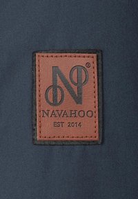 Brun lædermærke med præget logo "NAVAHOO" og "EST 2014," syet fast på en mørkeblå stofbaggrund.