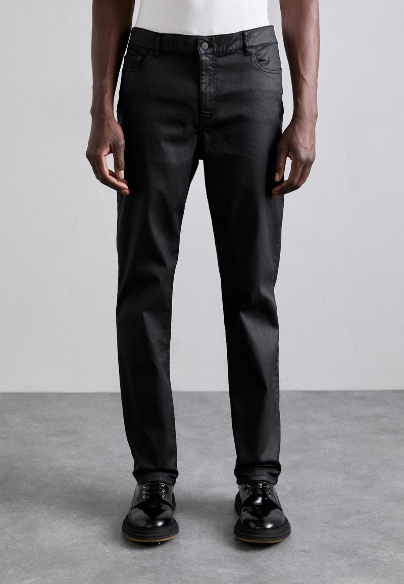 Pantalons noirs en denim, présentant une coupe slim, un design à cinq poches et une texture lisse, associés à des chaussures en cuir noires.