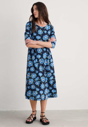 Floral dress in navy fabric featuring blue flowers with orange centers. Three-quarter sleeves, knee-length, paired with strappy black sandals.