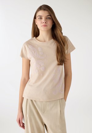 Giovane donna con capelli castani lunghi che indossa una t-shirt beige ricamata a maniche corte e pantaloni larghi beige chiaro, in piedi davanti a uno sfondo bianco.