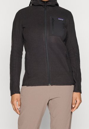 Personne portant une veste polaire Patagonia noire zippée avec une poche poitrine et un pantalon beige, debout devant un fond uni.