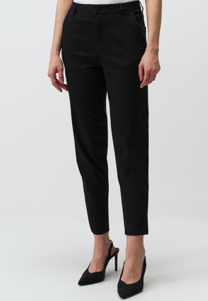 Pantaloni neri tapered in cotone, con tasche anteriori, chiusura con bottone e una texture liscia, abbinati a tacchi neri a punta.