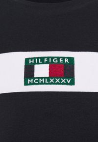 Černé bavlněné tričko s bílými horizontálními pruhy. Obsahuje vyšívané logo v zelené, bílé a červené barvě, kter�é zahrnuje nápisy "HILFIGER" a "MCMLXXXV."