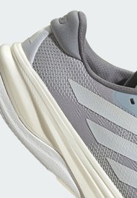 Baskets de sport grises avec un dessus en mesh texturé, un talon renforcé et une semelle blanche rembourrée ; comporte trois rayures gris clair sur le côté.