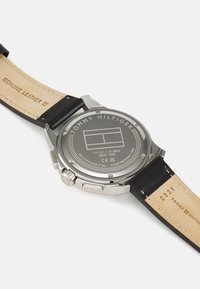 Reloj de acero inoxidable con una correa de cuero genuino negro. Presenta una parte trasera plateada grabada con "TOMMY HILFIGER" y detalles del modelo.