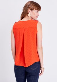 Top rosso senza maniche con retro plissettato, realizzato in tessuto leggero, con motivi delicati e spalline. Indossato con jeans scuri.