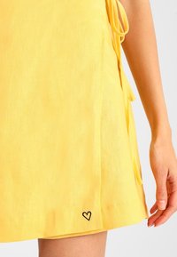 Gelbes Leinenkleid mit seitlichen Bändern und einem schwarzen Herz-Stickdetail nahe dem Saum. Glatte Textur mit lockerer Passform.