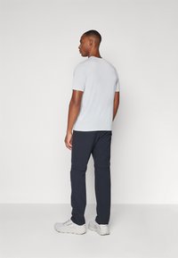 Chemise grise clair à manches courtes, pantalon bleu marine avec zips, et chaussures de sport blanches. Le modèle se tient de dos, avec un fond minimaliste.
