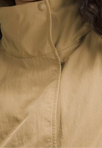 Gros plan sur le col d'un trench-coat beige avec des détails cousus et une section de cheveux ondulés foncés visible sur le côté droit.