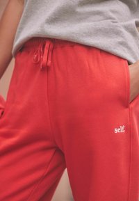 Red cotton sweatpants with elastic waistband and drawstring. Features side pockets and white logo text "self." on the left leg.