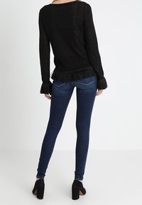 Černý dlouhý svetr s třásněmi na spodním lemu a na manžetách, v kombinaci s tmavomodrými skinny džínami a černými podpatkovými kotníkovými botami.