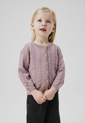 Gilet tricoté rose clair avec un motif en chevrons texturé, doté de boutons en bois et de manches longues, associé à un pantalon sombre.