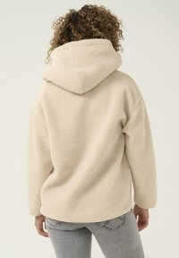 Jasno-kremowy polarowy hoodie o teksturowanej, miękkiej powierzchni i obszernym kapturze. Posiada luźne rękawy i swobodny krój.