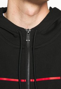 Sudadera negra con cremallera y textura suave, que presenta una cremallera central, capucha con cordón y dos líneas rojas horizontales en el pecho.