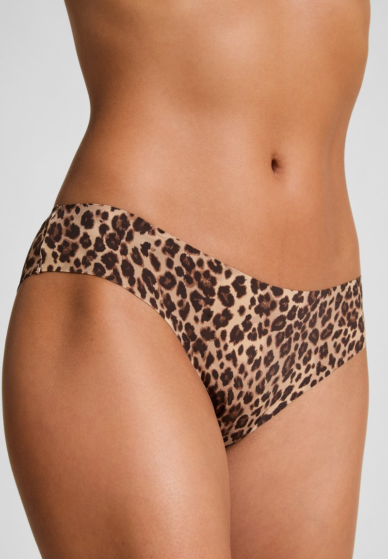Torse de femme portant un sous-vêtement hipster sans couture à motif léopard sur fond gris uni.