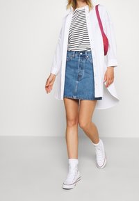 Kvinna som går och bär en randig skjorta, vit lång knappad skjorta, jeanskjol, vita höga sneakers och har en röd axelväska.