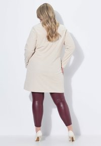 Donna con lunghi capelli biondi ondulati che indossa un vestito beige a maniche lunghe, leggings in pelle bordeaux e tacchi alti bianchi, girata di spalle contro un muro bianco.