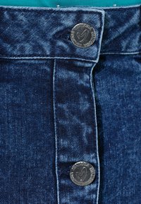 Tissu denim bleu foncé avec une texture lisse ; comprend deux boutons en métal à la taille pour la fermeture, affichant des gravures.