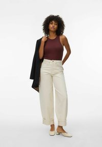 Femme aux cheveux bouclés portant un haut sans manches bordeaux, un pantalon large taille haute crème, des mules beiges, tenant une veste noire sur une épaule.