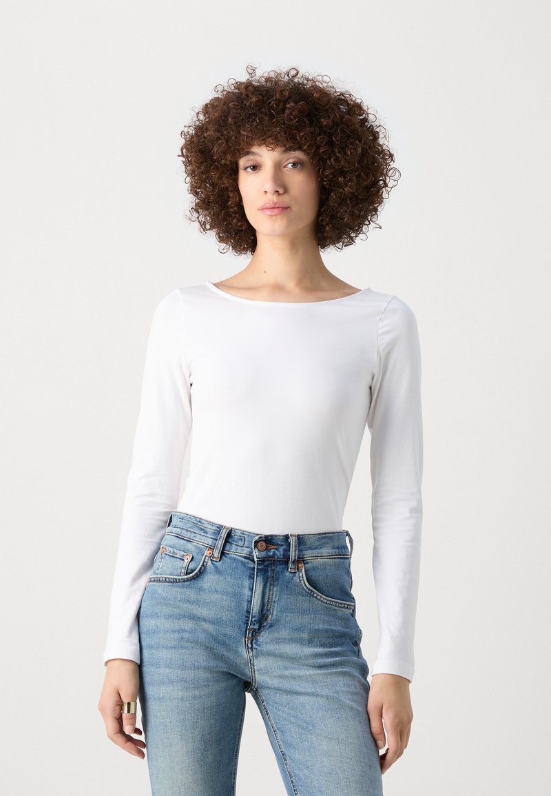 Haut blanc à manches longues avec un col bateau, associé à un jean bleu clair. Le haut présente une texture lisse et une coupe ajustée.