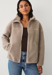 Veste en polaire beige clair avec un col montant, une texture douce, une fermeture zippée, et deux poches latérales. Présente des accents beige contrastants.
