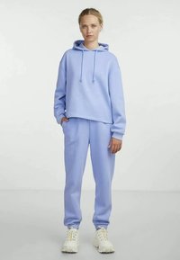 Hoodie azul claro com cordões, combinado com calças de treino azul claro. Ambas as peças têm uma textura de tecido suave e um design de ajuste solto.