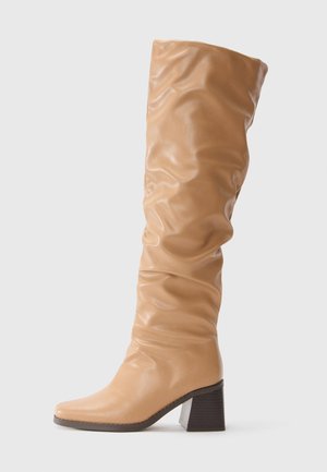 Botte en cuir beige haute avec un tige froissée et un talon carré,  bout pointu anguleux et semelle foncée sur fond clair.