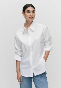 Weißes Baumwollhemd mit Knopfleiste, Kragen und hochgeschlagenen Ärmeln, im entspannten Schnitt. Kombiniert mit blauen Jeans.