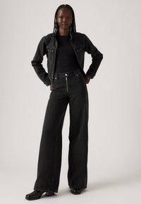 Giacca di jeans nera con due tasche frontali, abbinata a jeans neri a vita alta e gamba larga con zip anteriore e una leggera texture. Stivali neri.