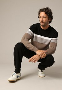 Gebreide trui met zwarte, witte en bruine horizontale strepen, gecombineerd met zwarte broek en witte sneakers. Model bevindt zich in een gehurkte positie.