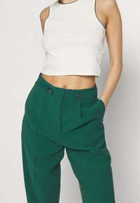 Bílý žebrovaný bezrukávový crop top s černým okrajem, který je spárován s vysokými zelenými kalhotami s předními kapsami a knoflíkovým uzavíráním.