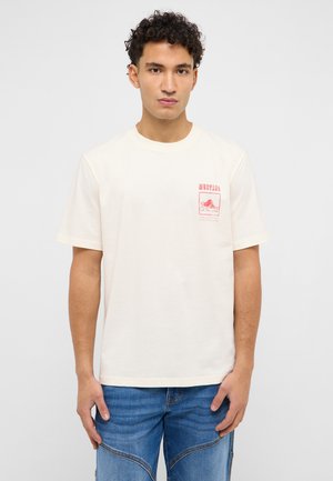 Junger Mann trägt ein cremeweißes T-Shirt mit Grafik und blaue Jeans, steht vor einem schlichten, hellgrauen Hintergrund.