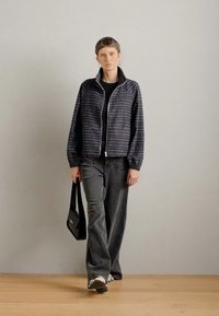 Πλαϊνί jacket με φερμουάρ σε σκούρους τόνους, σε συνδυασμό με γκρίζες παντελόνες wide-leg και μαύρη τσάντα ώμου, που στέκεται σε ξύλινο δάπεδο με ουδέτερο φόντο.