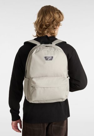 Mochila de tela beige con correas acolchadas y una asa superior. Cuenta con un compartimento central, un bolsillo frontal y un parche con el logo de Vans en la parte posterior.