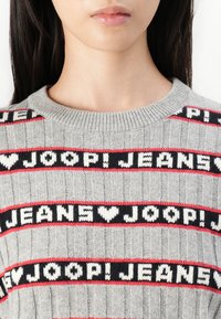 Maglione grigio lavorato a maglia con strisce orizzontali rosse e nere e testo bianco "JOOP! JEANS" e cuori. Collo a coste e texture visibile.