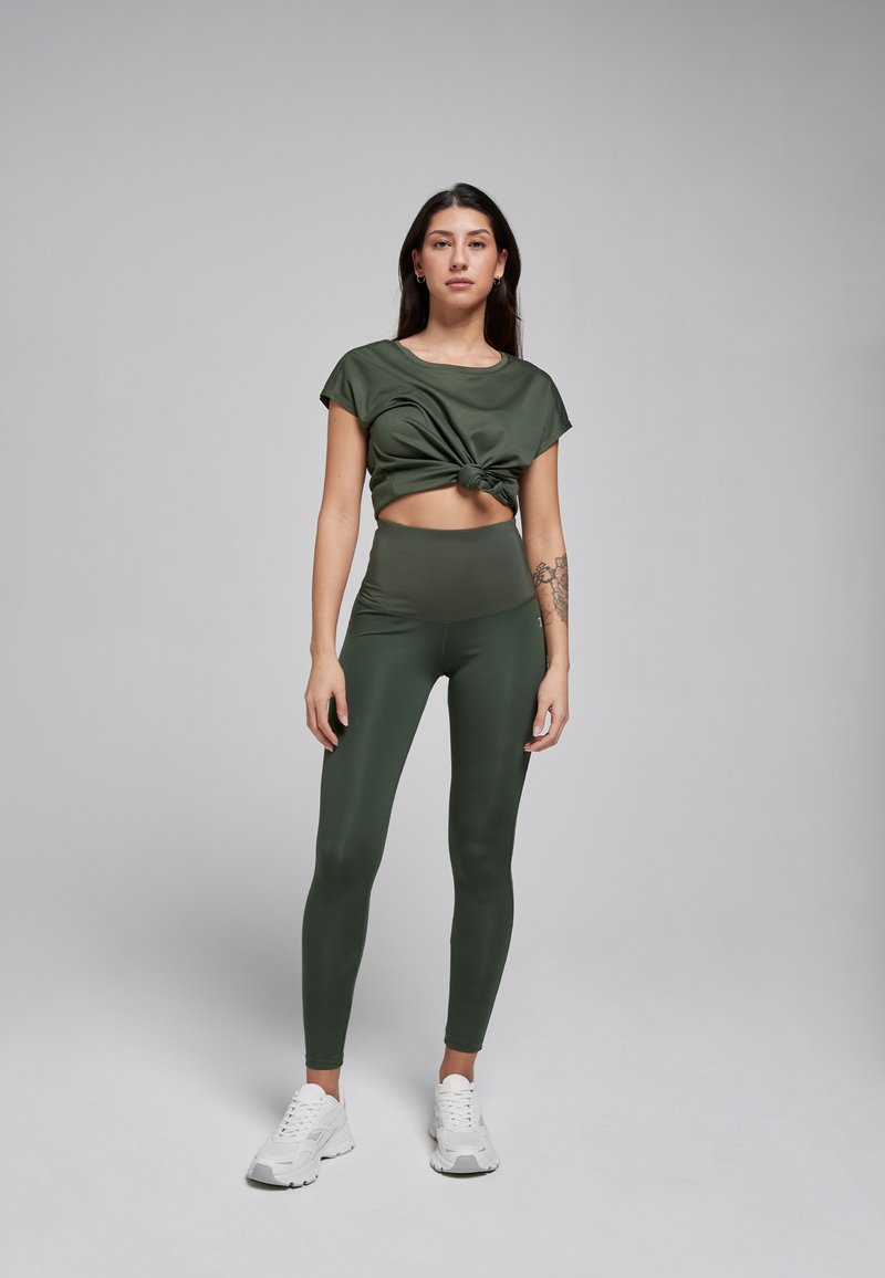 Olijfgroene sportset bestaande uit een knoop-detail crop top en hoge taille leggings, beiden vervaardigd uit een gladde, rekbare stof. Witte sneakers maken de look compleet.