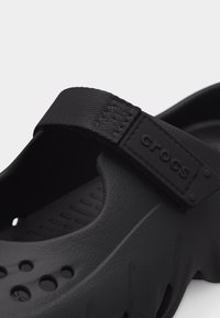 Sabot Crocs noir avec bride arrière en tissu texturé réglable et section supérieure ventilée perforée.