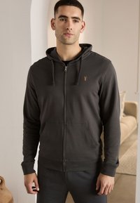 Pelēka zip-up džemperis ar kangarū maisiņu, auklītēm un mazu izsistam logotipu uz krūšu. Gluda kokvilnas auduma maisījums.