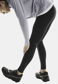 Leggings de sport noirs et chaussures de sport grises, présentant un tissu texturé et un logo sur le côté de la chaussure. L'utilisateur étire la jambe.