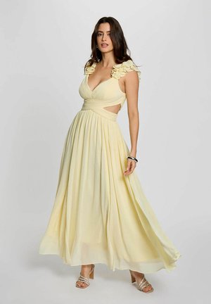 Donna che indossa un abito lungo giallo pallido senza maniche con spalline arruffate e lati con ritagli, in piedi davanti a uno sfondo chiaro e semplice.