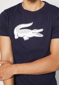 Marinblå t-shirt i bomull, med en vit krokodil-logotyp och triangulära mönster på kroppen. Korta ärmar och rundhalsad design.