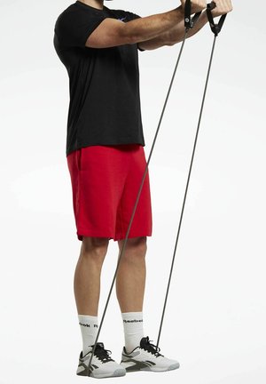 Man in zwart shirt en rode korte broek die met beide handen weerstandsbands omhoog trekt terwijl hij staat.