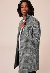 Cappotto di lana grigio a quadri con colletto alto, tasche laterali e vestibilità rilassata, indossato sopra una camicia bianca con un motivo testurizzato.
