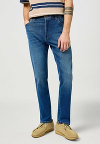 Jeans de mezclilla azul con corte recto, que cuentan con un estilo clásico de cinco bolsillos y un ligero desgaste. Llevados con zapatos tipo mocasín en tonos tostados.