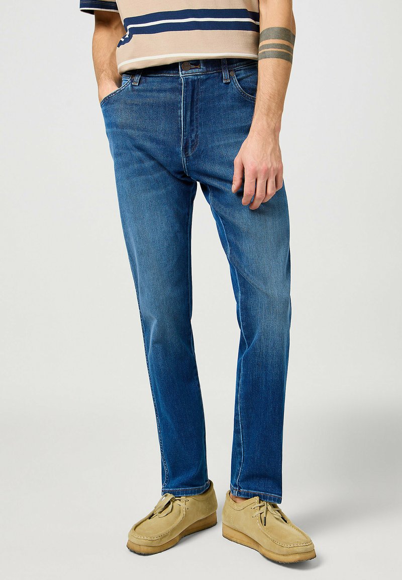 Jeans de mezclilla azul con corte recto, que cuentan con un estilo clásico de cinco bolsillos y un ligero desgaste. Llevados con zapatos tipo mocasín en tonos tostados.