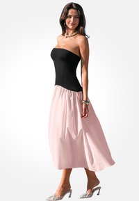 Zwarte strapless top met een luchtige lichtroze maxi-jurk. De jurk heeft een zachte, gestructureerde stof. Model draagt zilveren open teen schoenen.