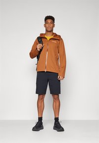 Brown zip-up jacket, yellow shirt, black shorts, carrying a black backpack. The jacket has a hood and front pocket; outfit suitable for outdoor activity.