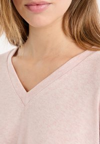 Sweter z dekoltem w serek w jasnoróżowym kolorze, wykonany z miękkiej tkaniny o delikatnej fakturze. Posiada luźny krój i prążkowany ściągacz przy dekolcie.